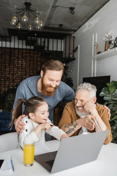 Smiling same sex parents doing homework with daughter near laptop at home — Stock Photo