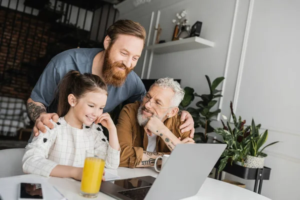 Smiling gay man hugging partner and daughter near devices and drinks at home — Stock Photo