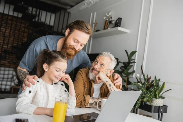 Sorridente uomo omosessuale abbracciando figlia e partner vicino a dispositivi e quaderni a casa — Foto stock