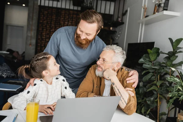 Overjoyed gay man hugging daughter and partner near laptop and orange juice at home — Stock Photo
