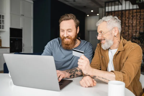 Carefree gay man holding credit card near partner with laptop at home — Stock Photo
