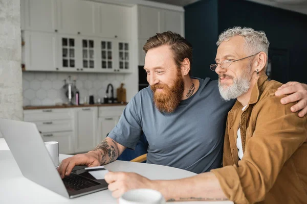 Усміхнений гомосексуальний чоловік використовує ноутбук і обіймає партнера з кредитною карткою вдома — стокове фото