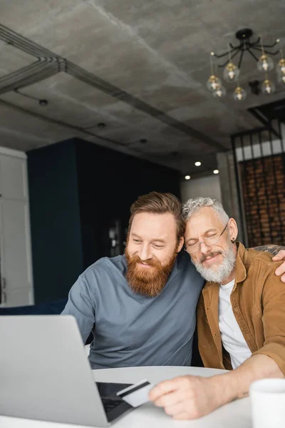 Carefree gay man using laptop and hugging partner with credit card near cup at home — Stock Photo