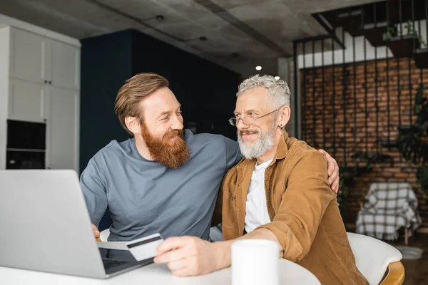 Positive gay man hugging partner with credit card near laptop and coffee at home — Stock Photo