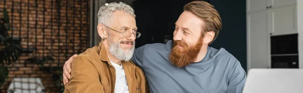 Alegre homosexual hombre abrazando madura pareja cerca borrosa portátil en casa, bandera - foto de stock