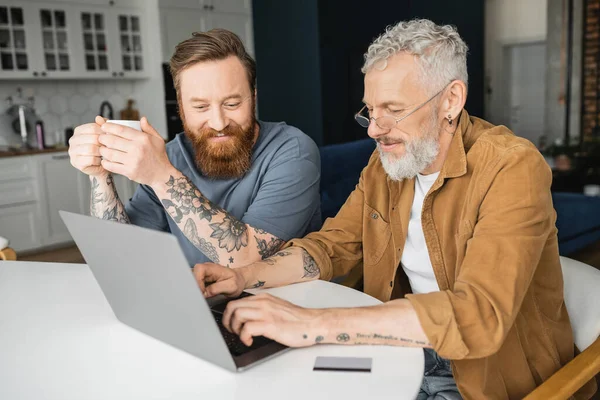 Smiling gay man holding cup of coffee while tattooed partner using laptop near credit card at home — Stock Photo