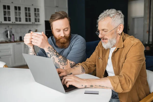Couple gay tatoué avec café à l'aide d'un ordinateur portable près de la carte de crédit à la maison — Photo de stock