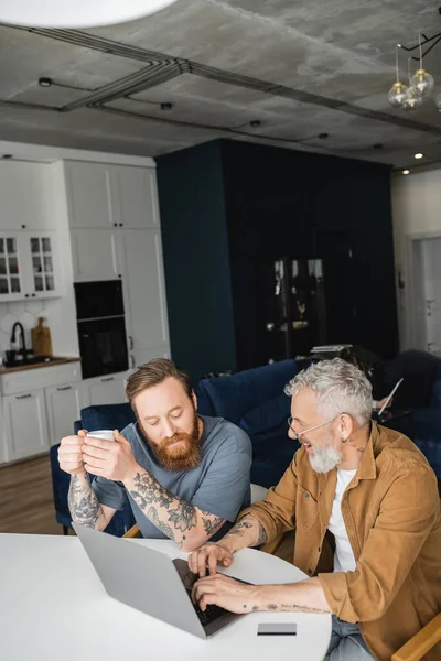 Tatuado gay homem segurando café e falando com parceiro usando laptop perto de cartão de crédito em casa — Fotografia de Stock