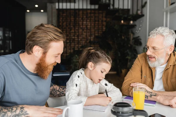 Tattooed gay fathers talking to daughter doing homework at home — Stock Photo
