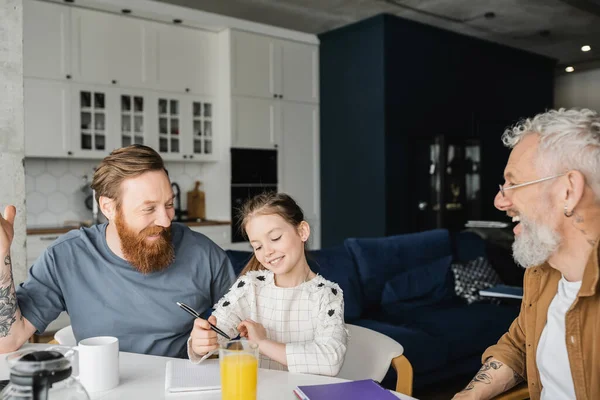 Smiling preteen girl doing homework near cheerful gay dads at home — Stock Photo