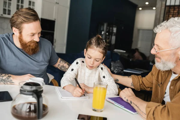 Smiling homosexual fathers looking at daughter doing homework at home — Stock Photo