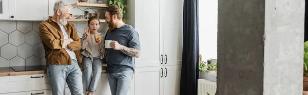 Same sex parents with coffee looking at daughter with croissant talking in kitchen, banner — Stock Photo