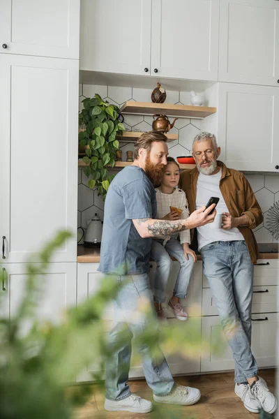 Homosexual father holding smartphone near daughter with croissant and partner in kitchen — Stock Photo