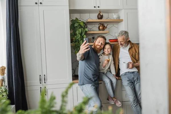 Smiling homosexual man taking selfie with daughter and partner in kitchen — Stock Photo