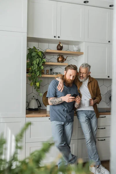 Sonriente gay hombre celebración taza y abrazo socio con celular en cocina - foto de stock