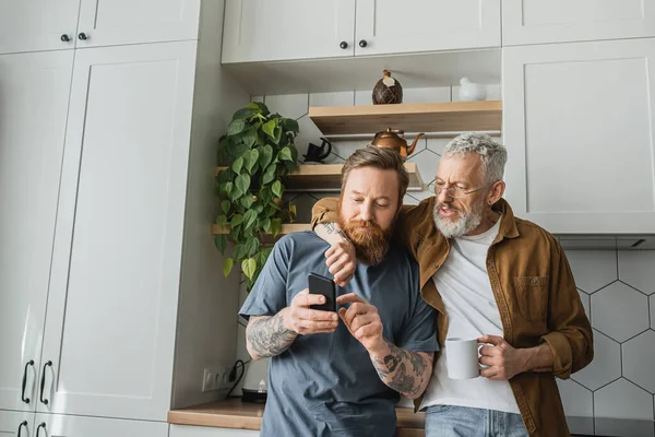 Gay hombre con taza de café abrazo socio con smartphone en cocina - foto de stock