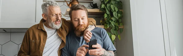 Homosexuel homme étreignant partenaire en utilisant un téléphone portable dans la cuisine, bannière — Photo de stock
