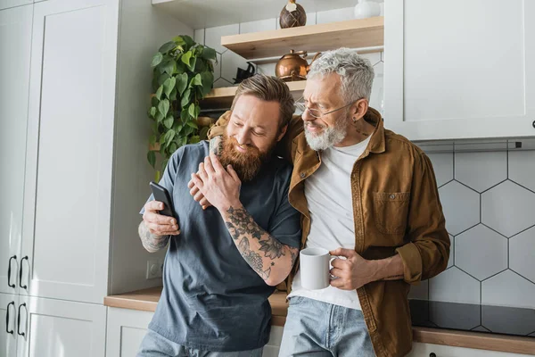 Hombre gay tatuado sosteniendo la taza y abrazando a su compañero sonriente con teléfono inteligente en la cocina - foto de stock