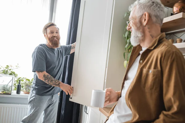 Cheerful tattooed gay man looking at blurred partner with coffee near cupboard in kitchen — Stock Photo
