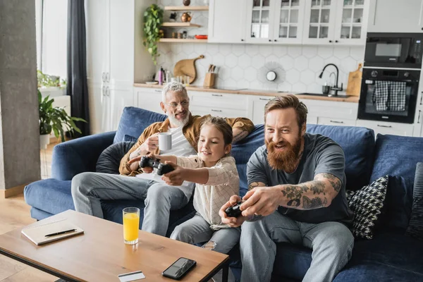 KYIV, UKRAINE - APRIL 19, 2023: Cheerful gay man playing video game with daughter near partner at home — Stock Photo