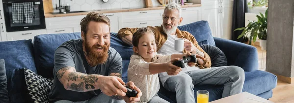 KYIV, UKRAINE - APRIL 19, 2023: Positive gay man playing video game with daughter near partner and orange juice at home, banner — Stock Photo