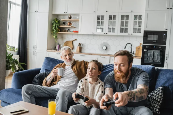 KYIV, UKRAINE - APRIL 19, 2023: Homosexual man playing video game with preteen daughter near partner at home — Stock Photo