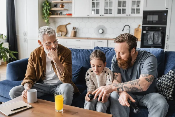 KYIV, UCRAINA - 19 APRILE 2023: Papà gay tatuato aiuta la figlia con joystick vicino al partner e beve a casa — Foto stock
