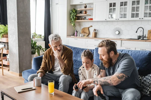 KYIV, UKRAINE - APRIL 19, 2023: Gay fathers helping daughter with joystick near drinks at home — Stock Photo
