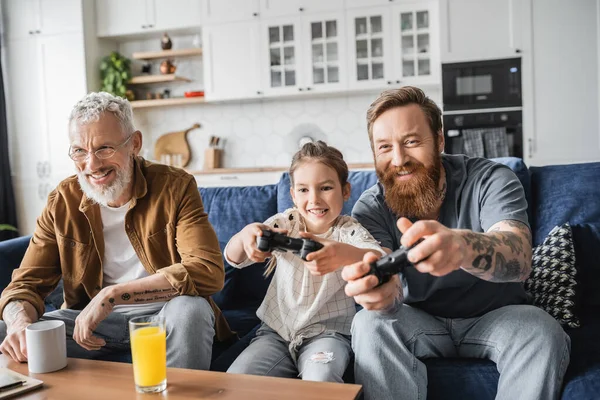 KYIV, UCRANIA - 19 DE ABRIL DE 2023: Sonriente padre gay jugando videojuego con su hija cercana a la pareja y bebiendo en casa - foto de stock