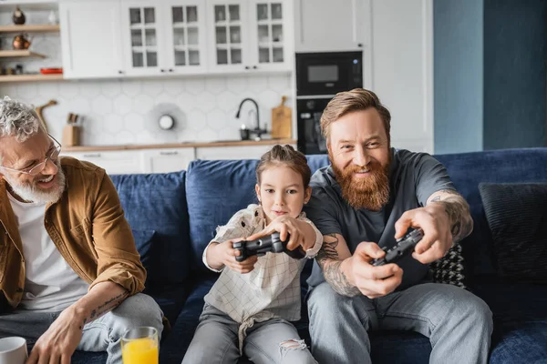 KYIV, UKRAINE - APRIL 19, 2023: Preteen girl playing video game near smiling homosexual dads at home — Stock Photo