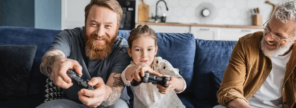 KYIV, UKRAINE - APRIL 19, 2023: Positive homosexual parents playing video game with daughter at home, banner — Stock Photo