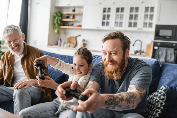 KYIV, UCRANIA - 19 de abril de 2023: Papá homosexual tatuado jugando videojuegos con una hija preadolescente cerca de su pareja en casa - foto de stock
