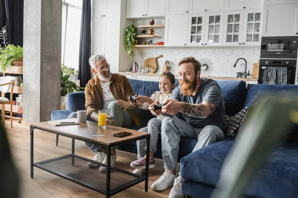 KYIV, UKRAINE - APRIL 19, 2023: Happy gay parents and preteen daughter playing video game on couch at home — Stock Photo