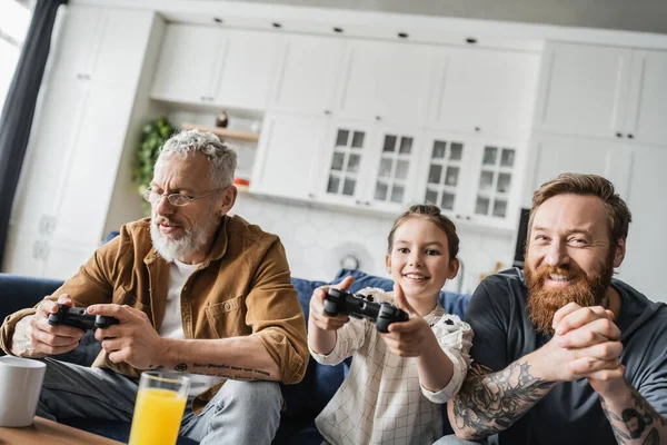KYIV, UKRAINE - APRIL 19, 2023: Cheerful kid playing video game near tattooed homosexual parents at home — Stock Photo