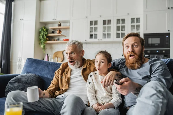 Shocked child watching tv near tattooed same sex parents at home — Stock Photo