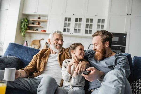 Smiling homosexual man with remote controller hugging daughter near partner with coffee at home — Stock Photo