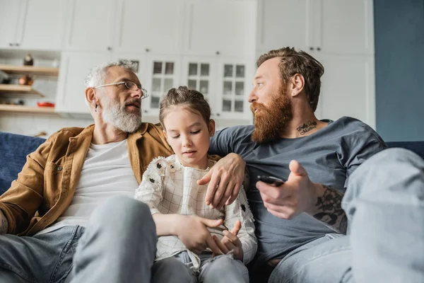 Padres gay con mando a distancia hablando cerca preadolescente hija en casa - foto de stock