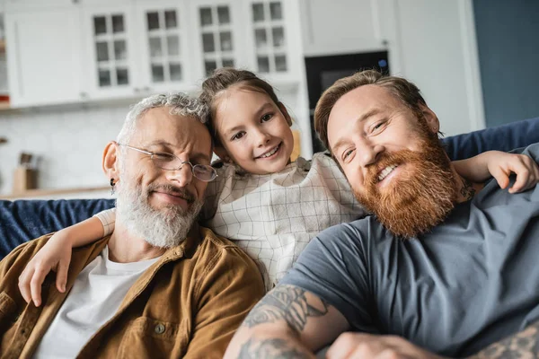 Sonriente niño abrazando barbudos padres homosexuales en el sofá en casa - foto de stock