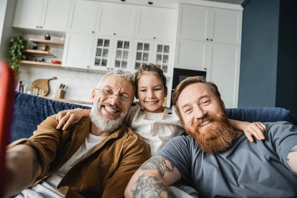 Lächelnde gleichgeschlechtliche Väter, die auf der heimischen Couch in die Kamera schauen — Stockfoto