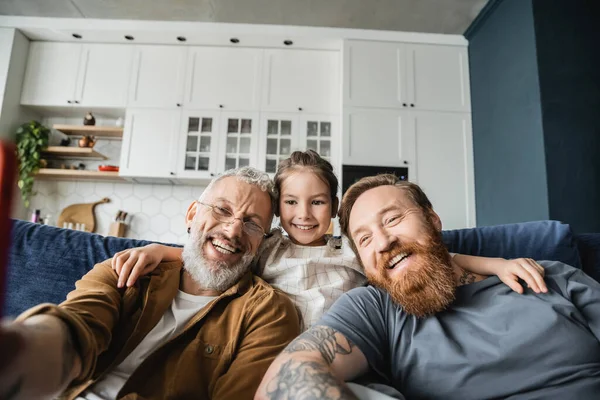 Беззаботная девушка обнимает бородатых гомосексуальных родителей на диване дома — стоковое фото