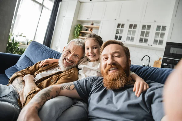 Sonriente niño preadolescente abrazando a los padres del mismo sexo y mirando a la cámara en el sofá en casa - foto de stock