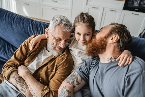 Vue grand angle de préadolescent souriant étreignant tatoué papas homosexuels sur le canapé à la maison — Photo de stock