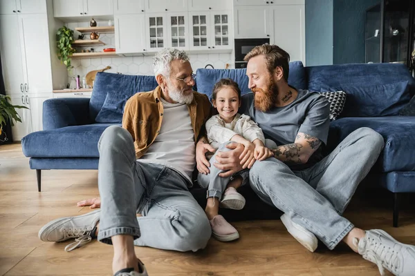 Tattooed homosexual parents looking at smiling daughter on floor in living room — Stock Photo