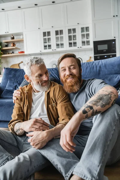 Positivo pareja del mismo sexo sentado cerca de sofá en el suelo en casa - foto de stock