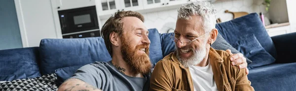 Татуированный гей обнимает веселого взрослого партнера в гостиной, баннер — стоковое фото