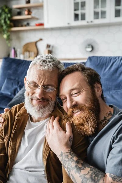 Татуированный и бородатый гей обнимает улыбающегося партнера дома — стоковое фото