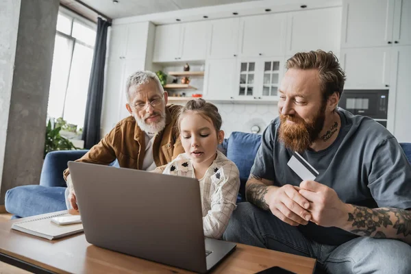 Preteen girl using laptop near smiling homosexual parent with credit card at home — Stock Photo