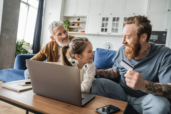 Padre homosexual sosteniendo tarjeta de crédito cerca de la hija sonriente con el ordenador portátil y la pareja en casa - foto de stock