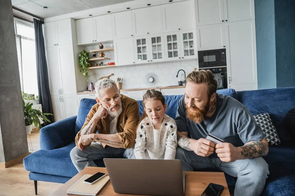 Schwule Eltern mit Kreditkarte schauen Tochter mit Laptop zu Hause an — Stockfoto