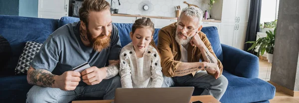 Preteen girl using laptop near tattooed same sex parents with credit card at home, banner — Stock Photo